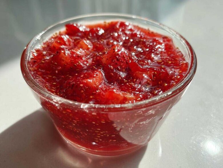 Close-up of vibrant red strawberry freezer jam with visible seeds, served in a small clear glass bowl.
