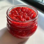 Close-up of a small glass jar filled with vibrant, chunky strawberry freezer jam, showing many seeds.