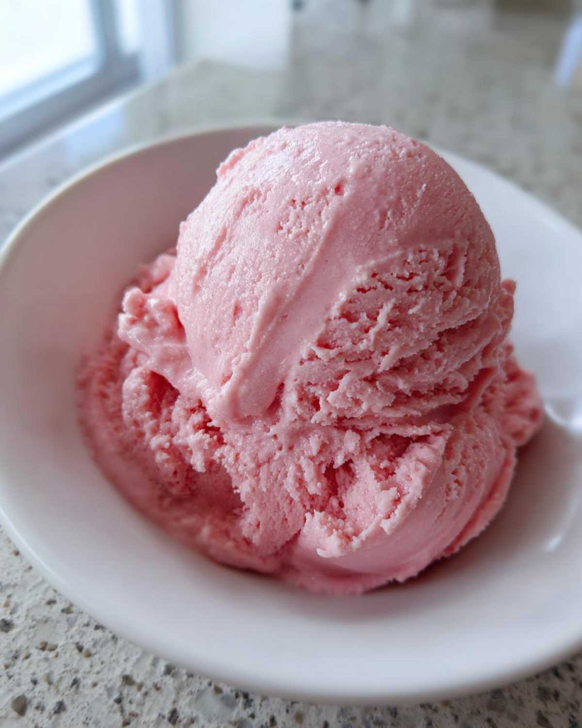 A close-up of a generous scoop of bright pink strawberry ice cream served in a white bowl.