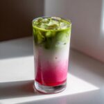 A tall glass showing a layered strawberry matcha drink with a bright pink bottom layer, white milk layer, and green matcha top layer over ice.