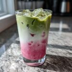 A tall glass featuring a layered strawberry matcha drink with bright pink puree, white milk, and green matcha tea over ice.