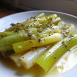 Close-up of tender braised leeks, glistening in butter or sauce and topped with fresh thyme.