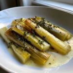 Close-up of tender braised leeks served in a white bowl with butter sauce and fresh thyme.