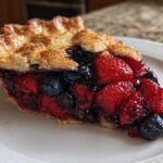 A close-up of a generous slice of triple berry pie showing juicy strawberries, blueberries, and a flaky golden crust.