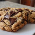 Three freshly baked vegan chocolate chip cookies with gooey, melted chocolate chunks on a white plate.