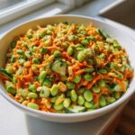 Close-up of a white bowl filled with bright green edamame salad mixed with shredded carrots and cucumber pieces.