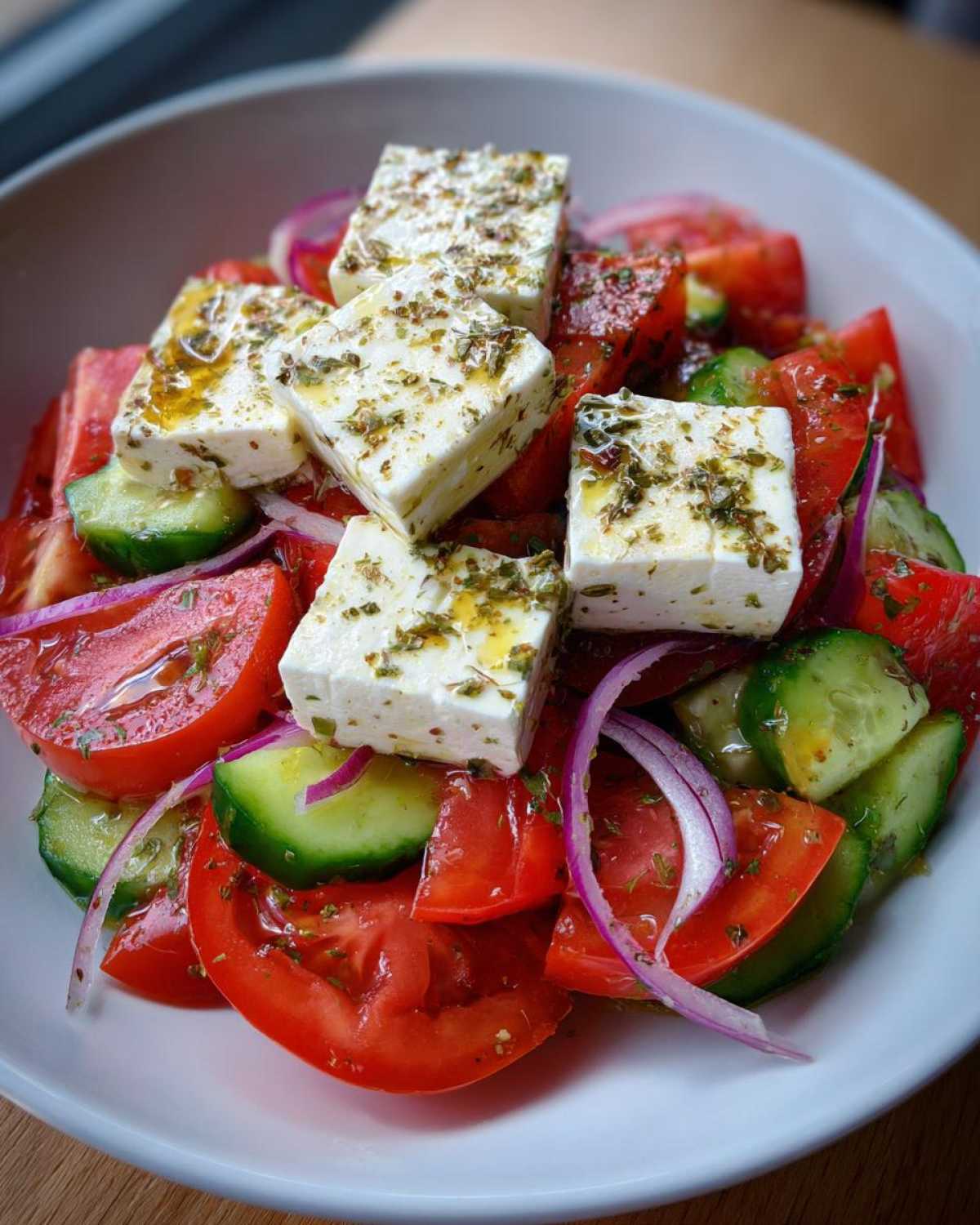 Close-up of a vibrant greek salad featuring large cubes of feta cheese drizzled with olive oil and herbs, mixed with tomatoes, cucumbers, and red onion.