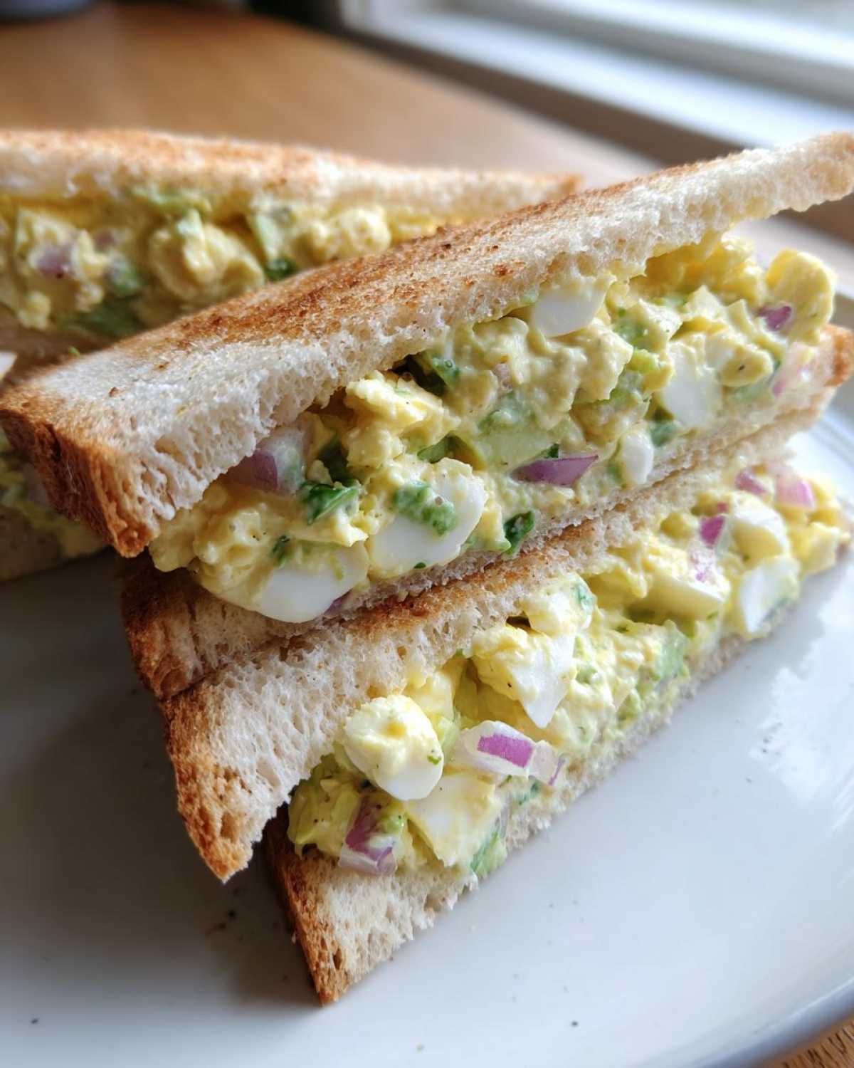 Two halves of an Avocado Egg Salad Sandwich stacked on a white plate, showing creamy filling with egg chunks and red onion.