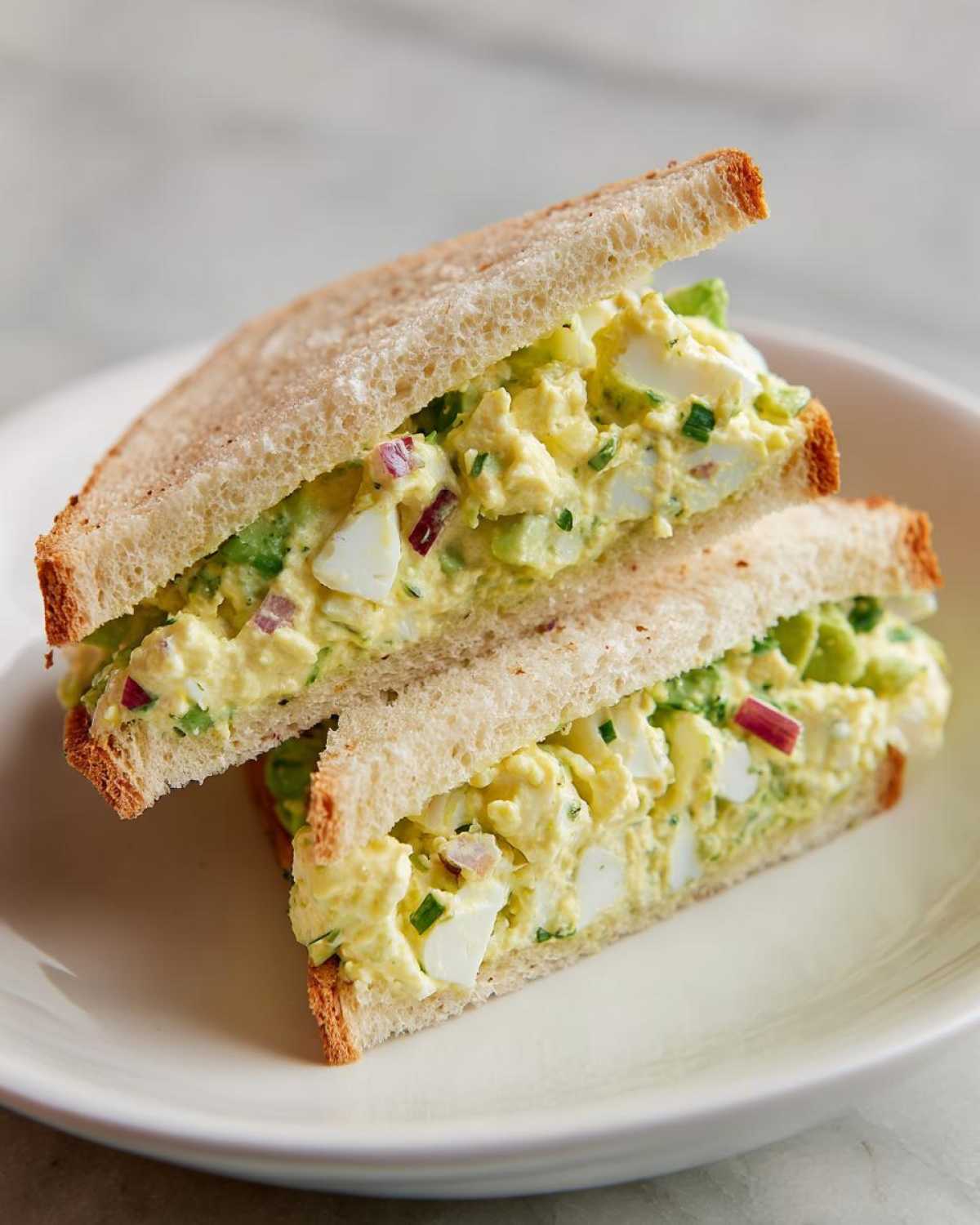 Two halves of an Avocado Egg Salad Sandwich stacked on a white plate, showing creamy filling with egg chunks and red onion.