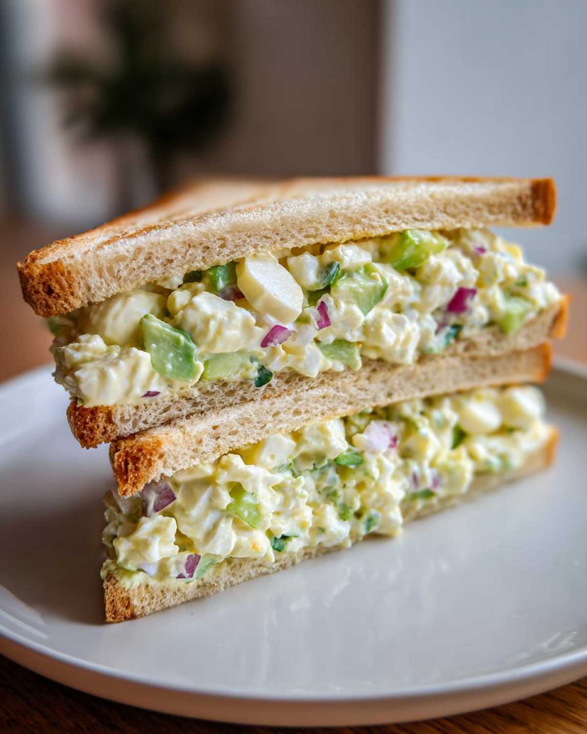 Two halves of an Avocado Egg Salad Sandwich stacked on a white plate, showing creamy filling with avocado chunks and red onion.