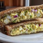 A hearty Avocado Egg Salad Sandwich, cut in half, showing creamy filling with egg chunks and red onion on toasted whole wheat bread.