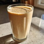 A tall glass filled with a thick, creamy Banana Peanut Butter Smoothie sitting on a speckled countertop in bright sunlight.