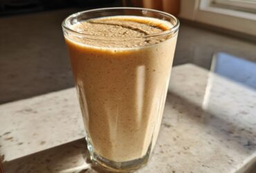 A tall glass filled with a thick, creamy Banana Peanut Butter Smoothie sitting on a speckled countertop in bright sunlight.