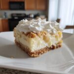 A single serving of Banana Pudding Cheesecake Bars topped with whipped cream and crumbs on a white plate.