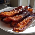 Close-up of crispy, shiny Brown Sugar Glazed Bacon strips resting on a paper towel on a white plate.