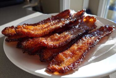 Close-up of crispy, shiny Brown Sugar Glazed Bacon strips resting on a paper towel on a white plate.