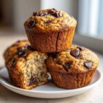 A stack of three golden-brown Chocolate Chip Banana Muffins, one broken open to show the moist interior.