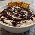 A white bowl filled with creamy Chocolate Peanut Butter Cheesecake Dip, drizzled with fudge and topped with chocolate shavings, served with crackers.