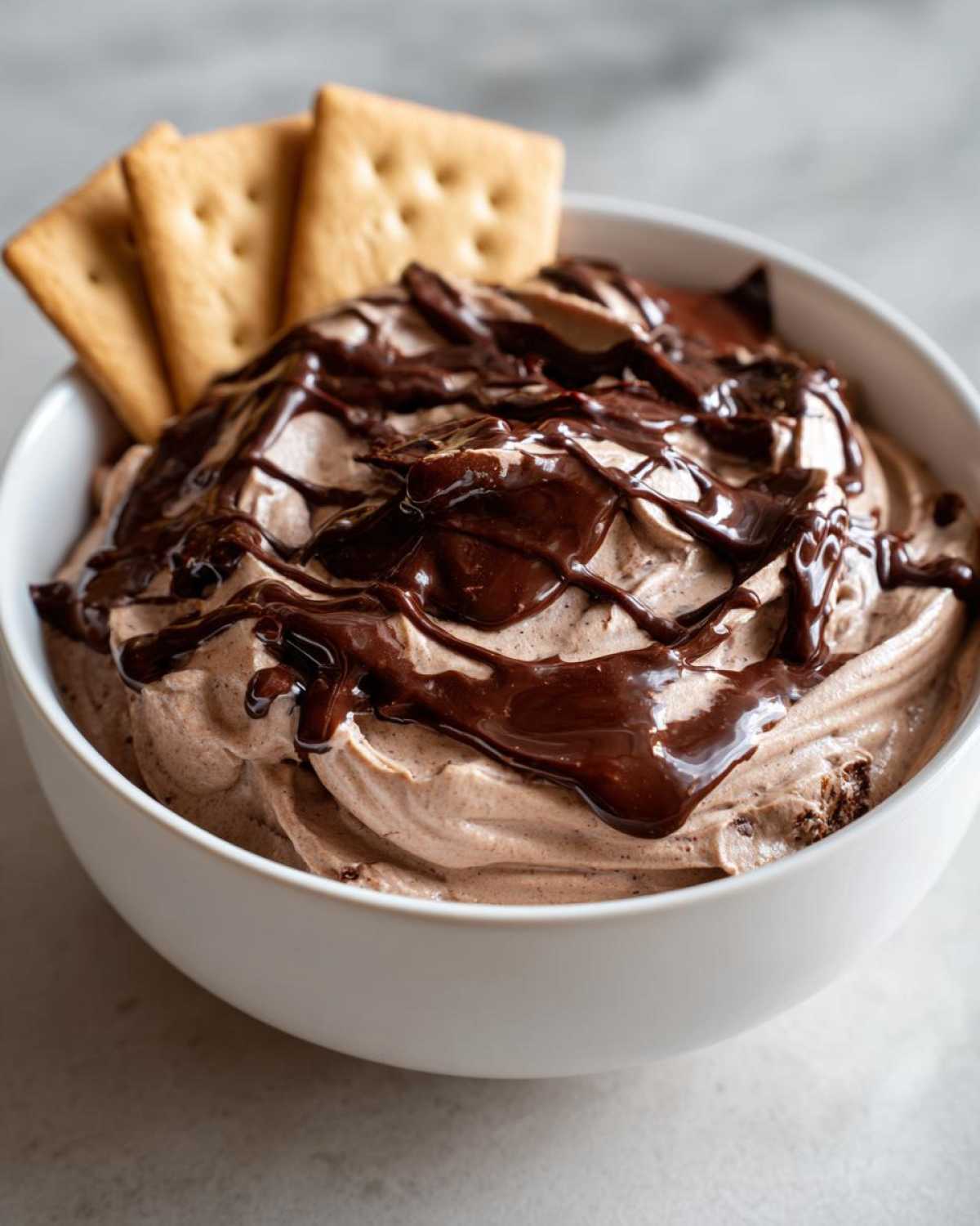 A bowl of creamy Chocolate Peanut Butter Cheesecake Dip topped with chocolate syrup and served with crackers.