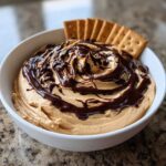 A white bowl filled with creamy Chocolate Peanut Butter Cheesecake Dip, topped with chocolate syrup and served with graham crackers.
