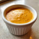 Close-up of thick, orange Copycat Raising Cane’s Sauce speckled with pepper in a small white fluted ramekin.
