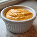 A close-up of thick, orange-yellow Copycat Raising Cane’s Sauce speckled with red spices in a small white ramekin.