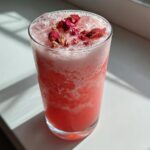 A tall glass filled with a vibrant pink drink, ice, foam, and topped with dried strawberry pieces, resembling a Copycat Starbucks Pink Drink.