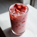 A tall glass filled with the pink, creamy Copycat Starbucks Pink Drink, topped with ice and fresh strawberry slices.