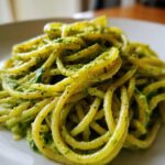 A close-up shot of thick spaghetti coated in bright green, creamy avocado pasta sauce, served on a white plate.