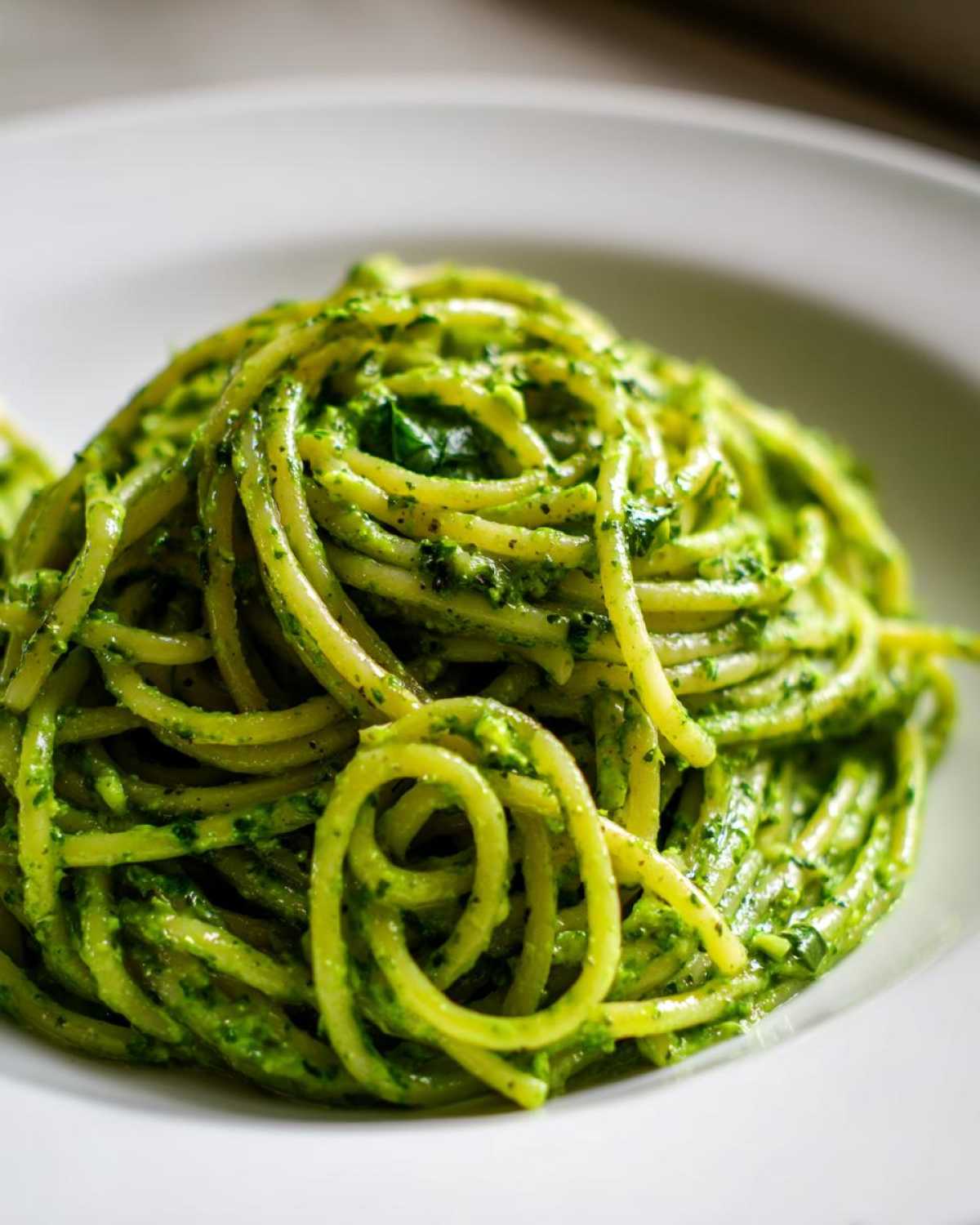 A close-up shot of spaghetti coated in vibrant green, creamy avocado pasta sauce, served on a white plate.