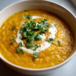 Close-up of a bowl of vibrant orange Creamy Coconut Lentil Soup, topped with coconut cream swirl and fresh cilantro.