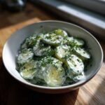 A close-up of a bowl filled with slices of Creamy German Cucumber Salad coated in a white dressing and topped with fresh dill and chives.
