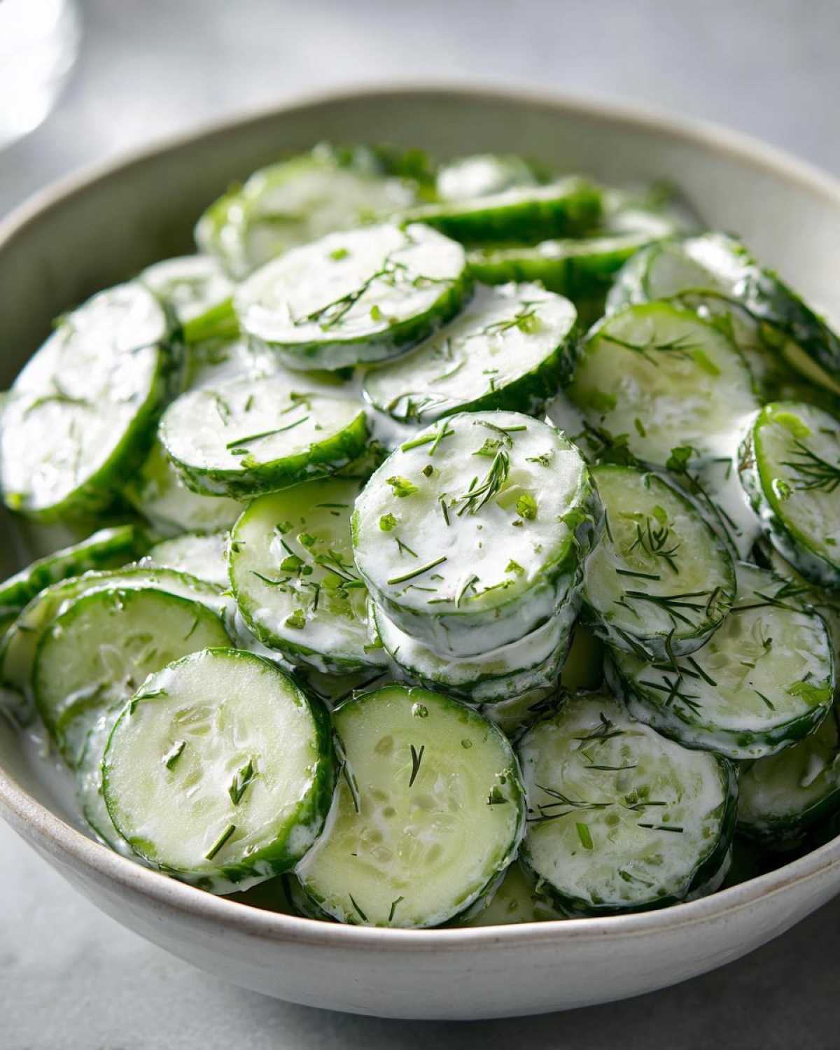 A bowl filled with thinly sliced cucumbers coated in a creamy dressing and topped with fresh dill, showcasing the Creamy German Cucumber Salad.