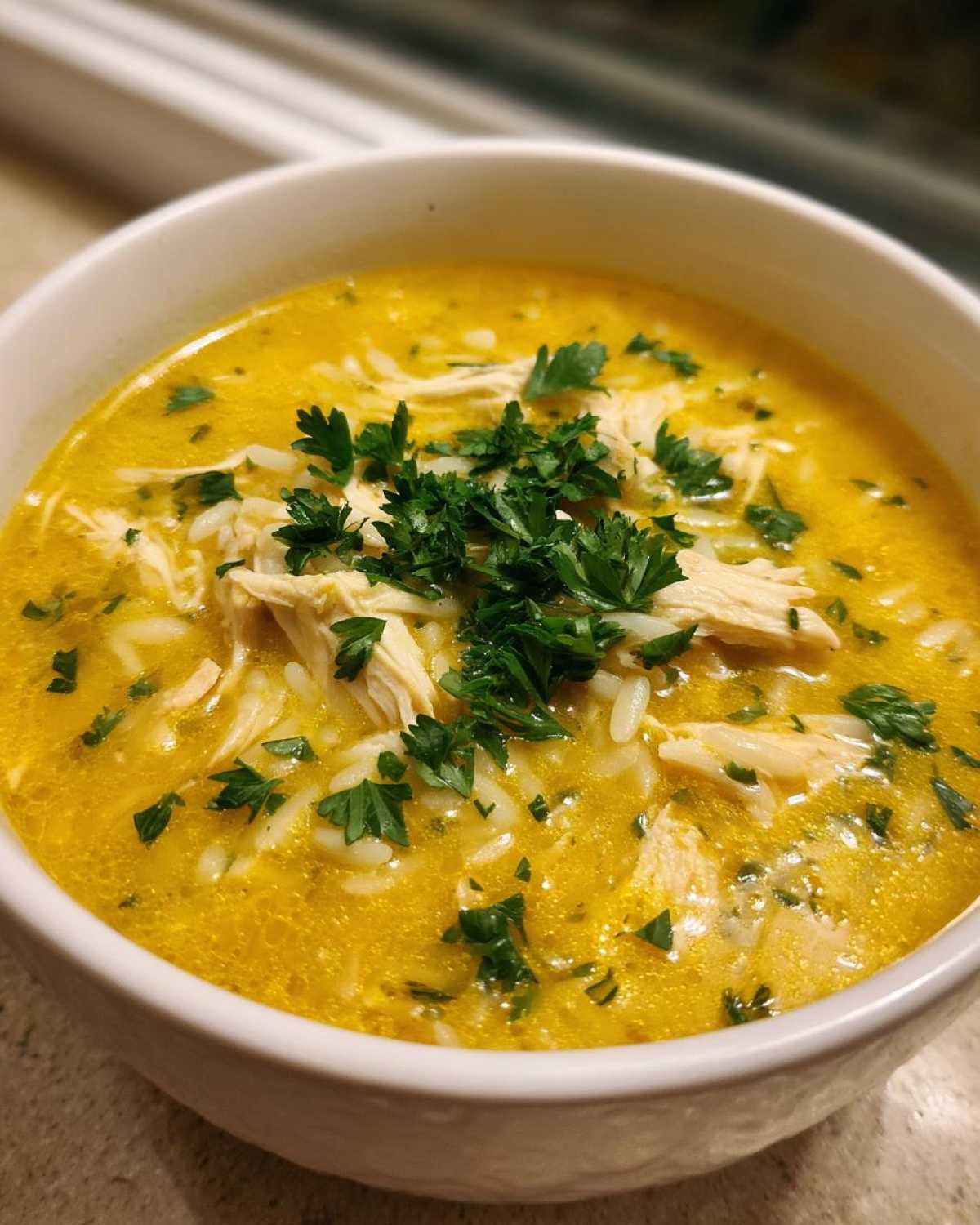 Close-up of a white bowl filled with bright yellow Creamy Lemon Chicken Orzo Soup, topped with shredded chicken and fresh parsley.