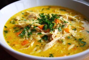 A close-up view of a bowl filled with rich, yellow Creamy Lemon Chicken Orzo Soup, topped with shredded chicken, carrots, and fresh parsley.