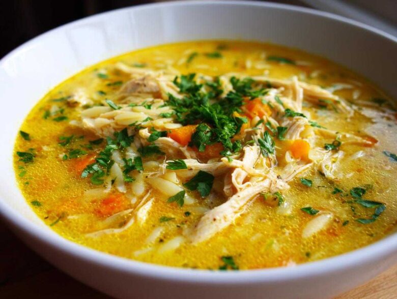 A close-up view of a bowl filled with rich, yellow Creamy Lemon Chicken Orzo Soup, topped with shredded chicken, carrots, and fresh parsley.