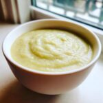 Close-up of a white bowl filled with smooth, creamy potato leek soup, set near a window.