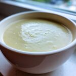 Close-up of a white bowl filled with thick, smooth Creamy Potato Leek Soup, set near a window.