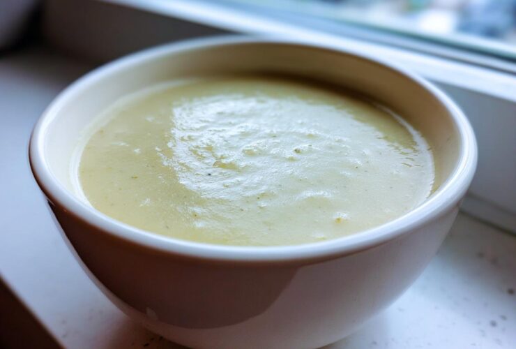 Close-up of a white bowl filled with thick, smooth Creamy Potato Leek Soup, set near a window.