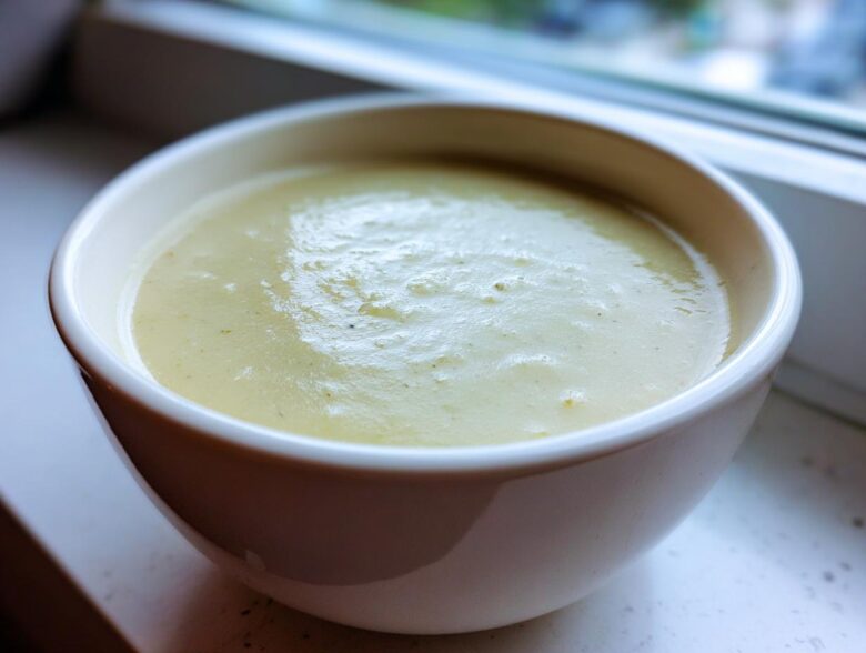 Close-up of a white bowl filled with thick, smooth Creamy Potato Leek Soup, set near a window.