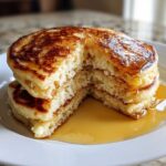 Close-up of a stack of fluffy Gluten-Free Buttermilk Pancakes cut open, drizzled generously with maple syrup.