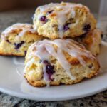 Close-up of several glazed Blueberry Lemon Scones stacked on a white plate, showing visible blueberries.