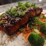Close-up of a glazed Teriyaki Salmon fillet served over white rice with broccoli, shredded carrots, and green onions.