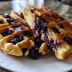 Close-up of fluffy Gluten-Free Blueberry Waffles, one piece cut open showing juicy blueberries inside, drizzled with syrup.