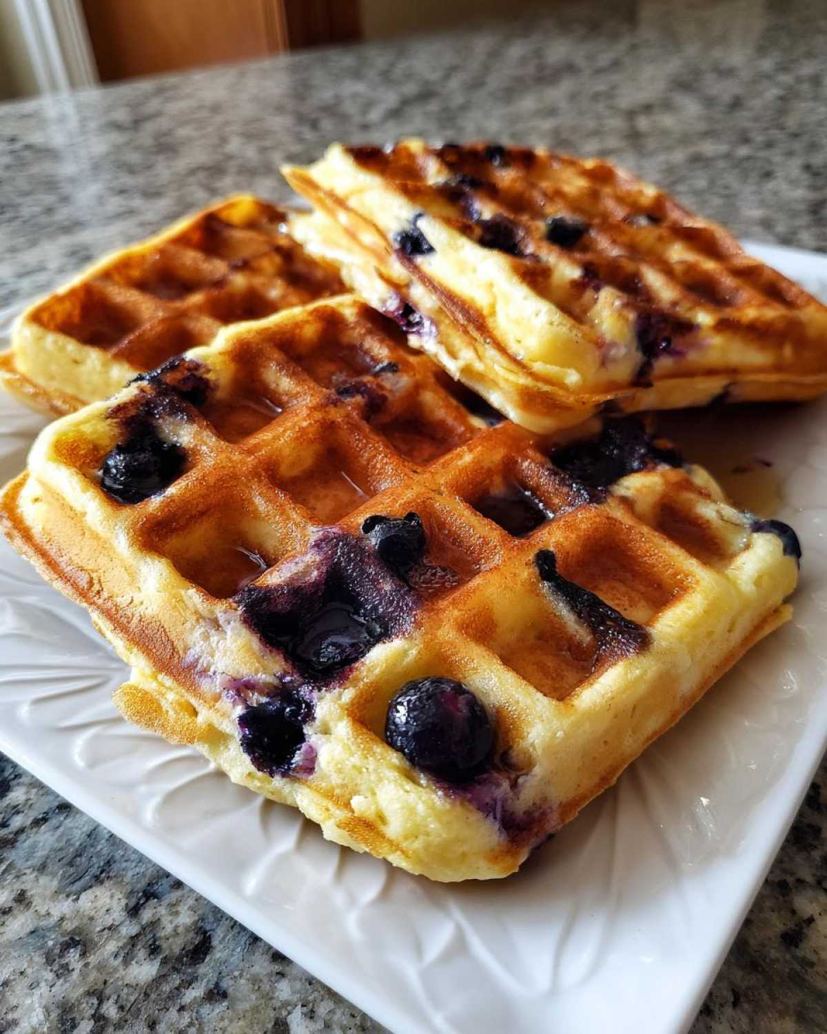 Close-up of three golden Gluten-Free Blueberry Waffles drizzled with syrup on a white plate.