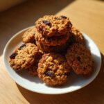A stack of freshly baked Gluten-Free Oatmeal Raisin Cookies piled on a white plate, highlighted by natural sunlight.