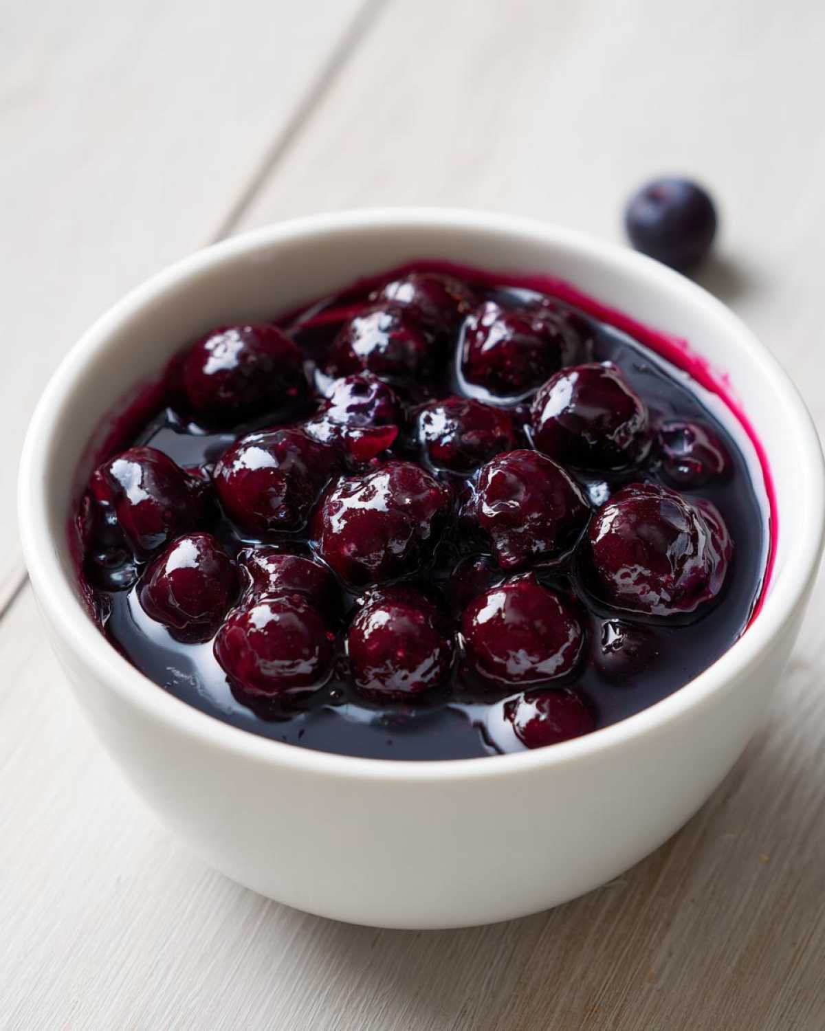 A close-up of plump blueberries simmering in thick, dark purple liquid, representing Homemade Blueberry Syrup, in a white bowl.