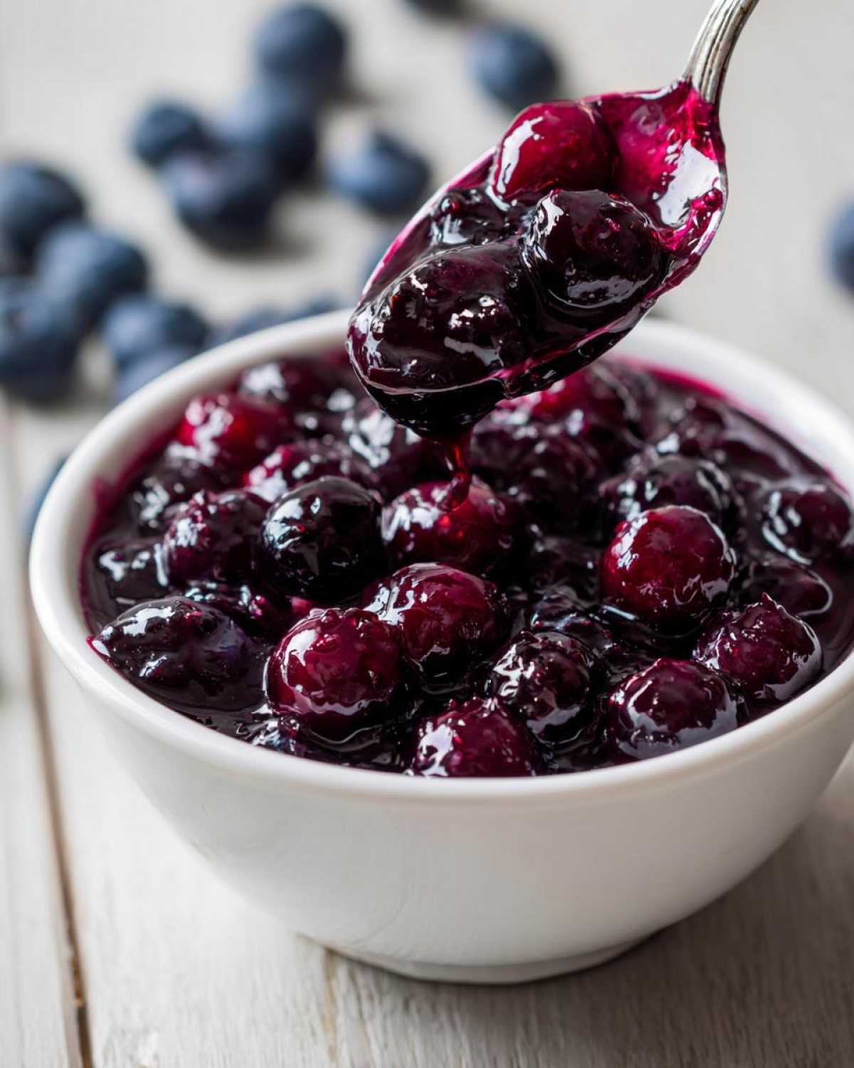A spoonful of thick, rich Homemade Blueberry Syrup being lifted from a white bowl filled with whole blueberries.