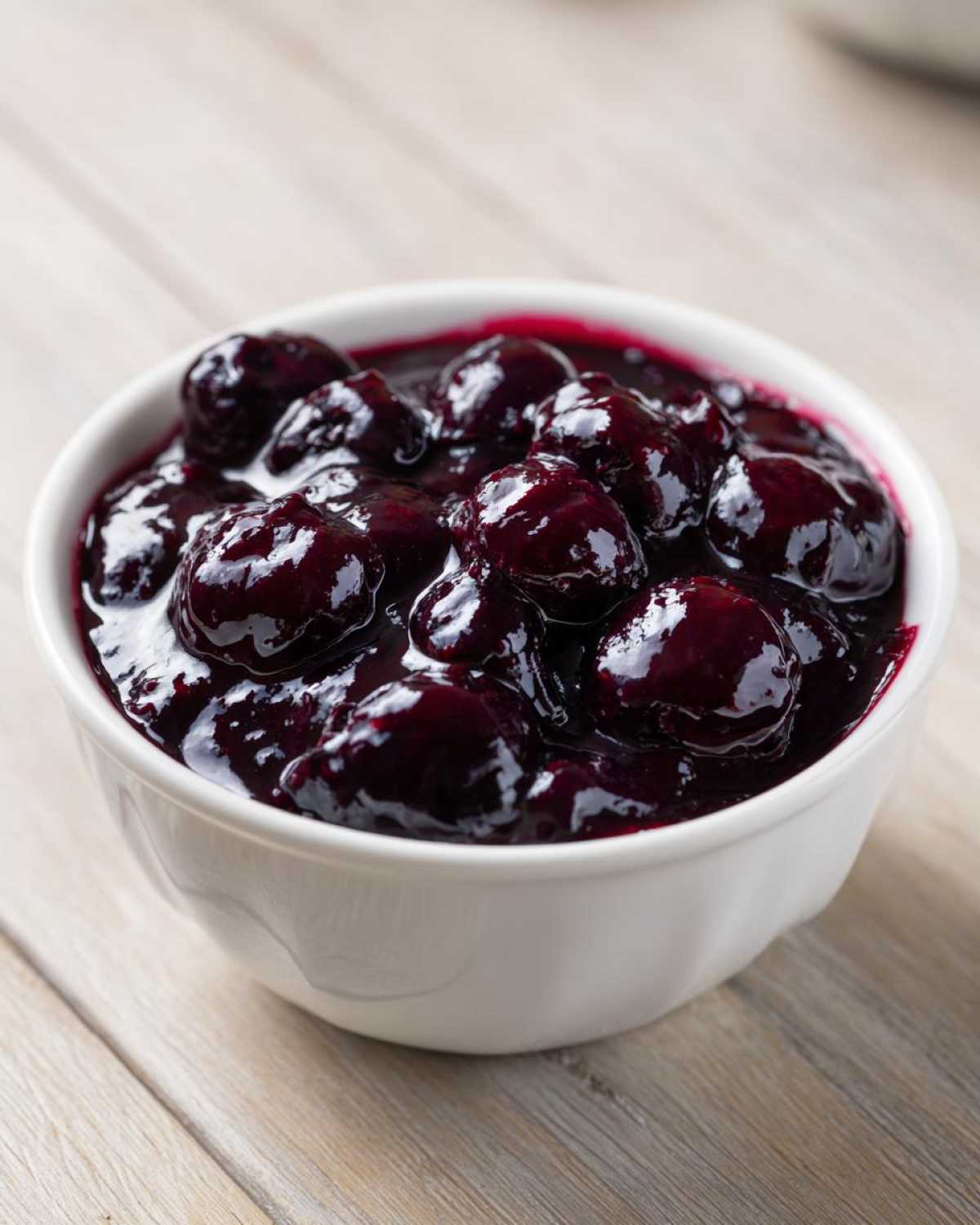 Close-up of rich, dark purple Homemade Blueberry Syrup topping with whole berries in a white bowl.