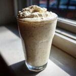 A tall glass filled with a speckled Homemade Vanilla Bean Frappuccino topped with whipped cream, backlit by a window.